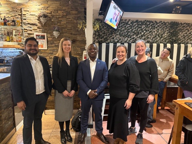 Five people—three women and two men—are standing and posing for a photo in a casual restaurant or cafe setting. A man in a blue suit is at the center, flanked by four colleagues.