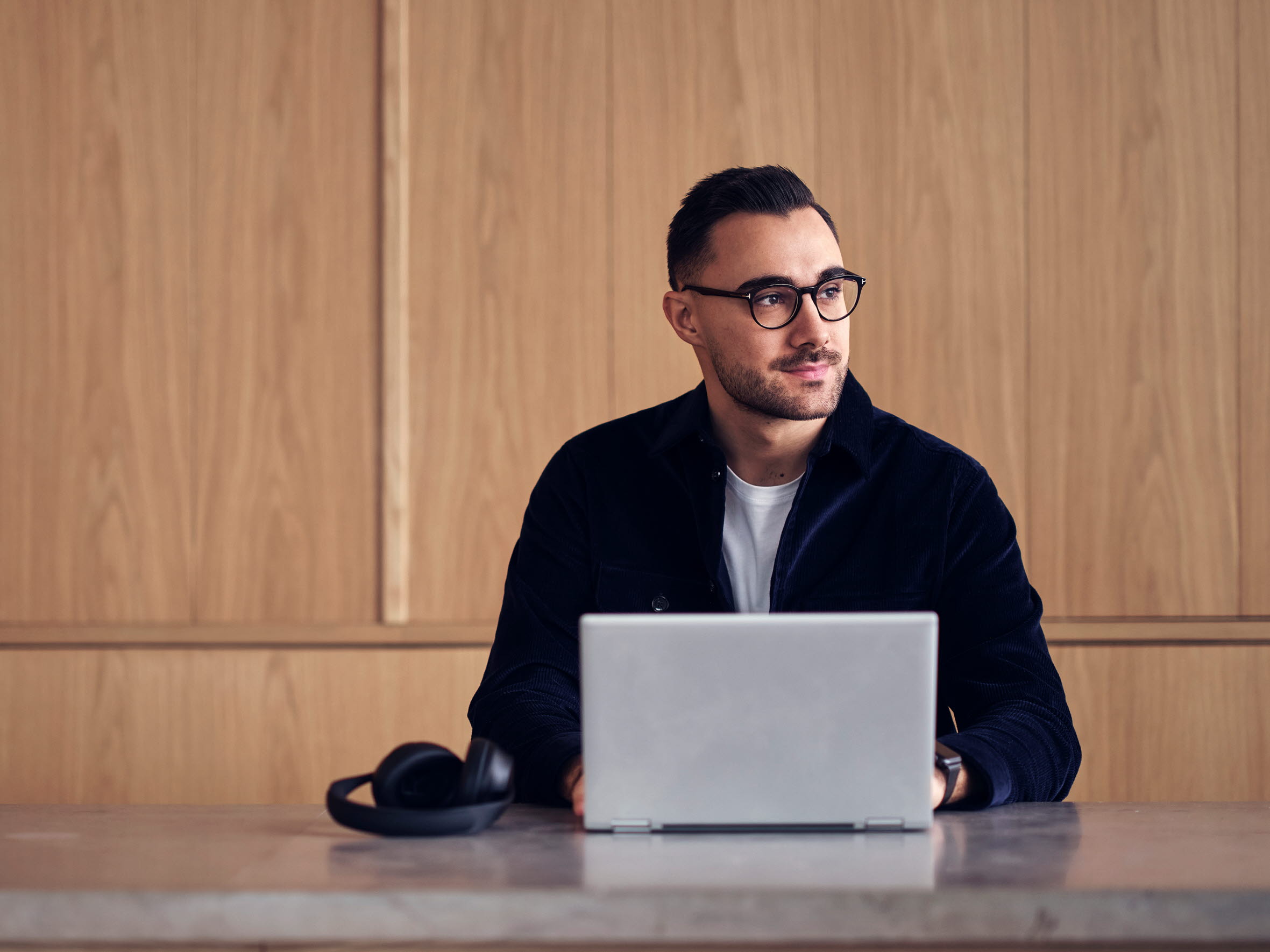 En ung man med glasögon som sitter vid ett skrivbord med sin laptop och jobbar.