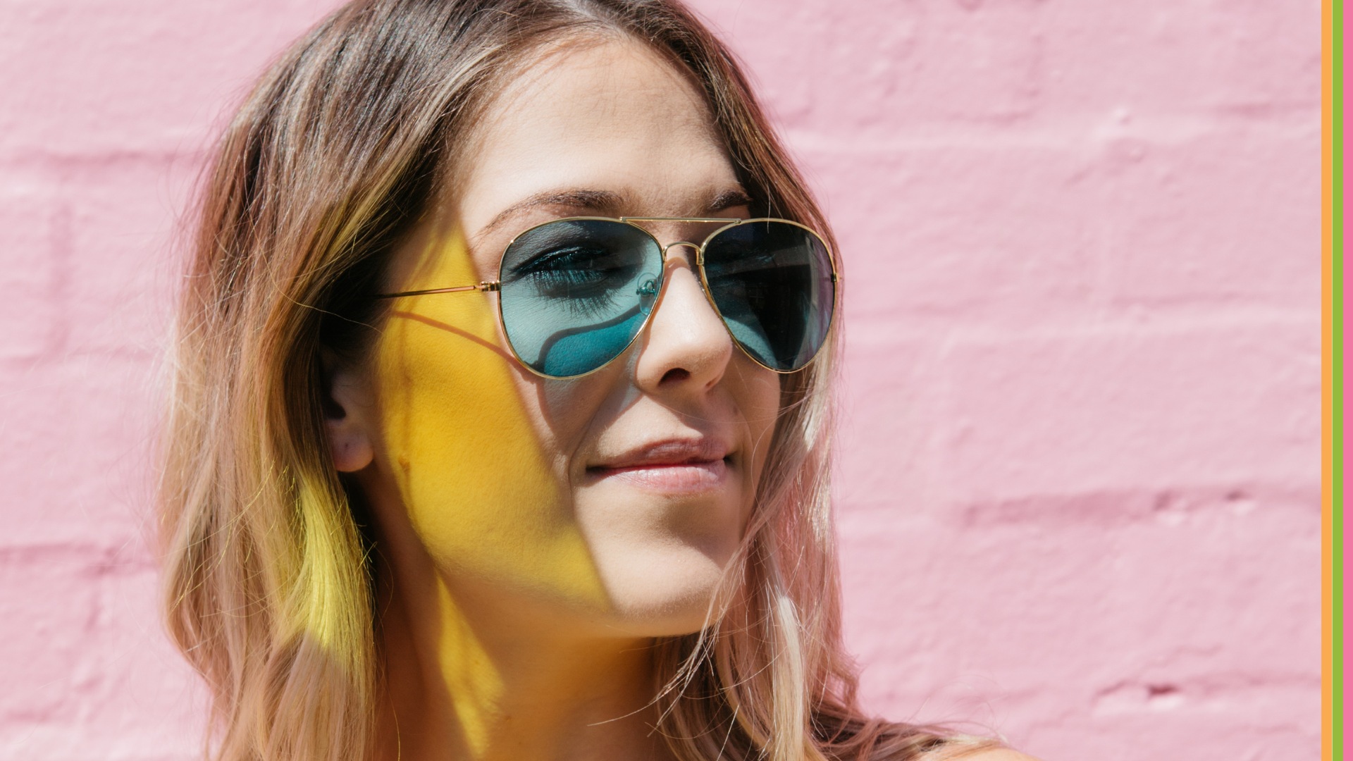 Woman with blonde hair wearing sunglasses with blue lenses, with yellow light shadow striking across face, in front of pink painted brick wall.