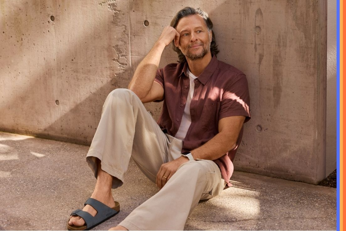 A man in a maroon shirt, beige pants, and dark Birkenstock sandals sits on the ground outdoors against a concrete wall.