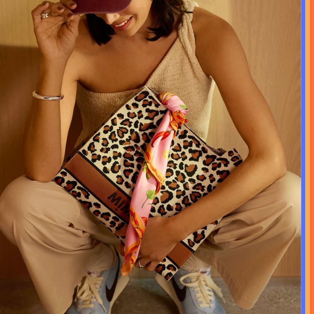 Woman holding a large leopard print handbag with a colorful scarf tied to the handle, styled with a sleeveless yellow top, beige pants, and light blue Nike sneakers.