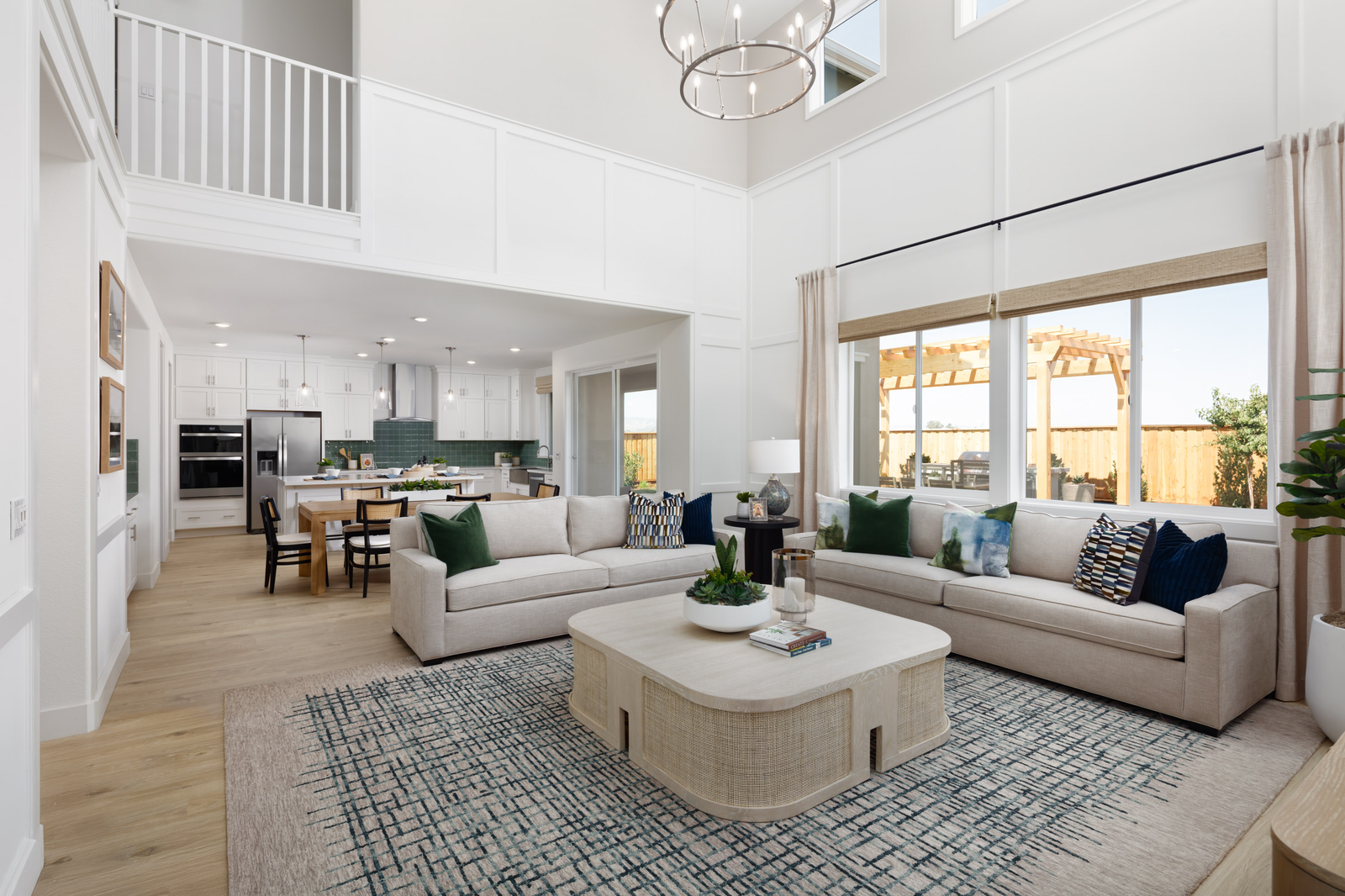 great room with high volume ceiling, wood floors, two tan sofas with green & blue pillows, light wood coffee table, and large windows looking out to the backyard