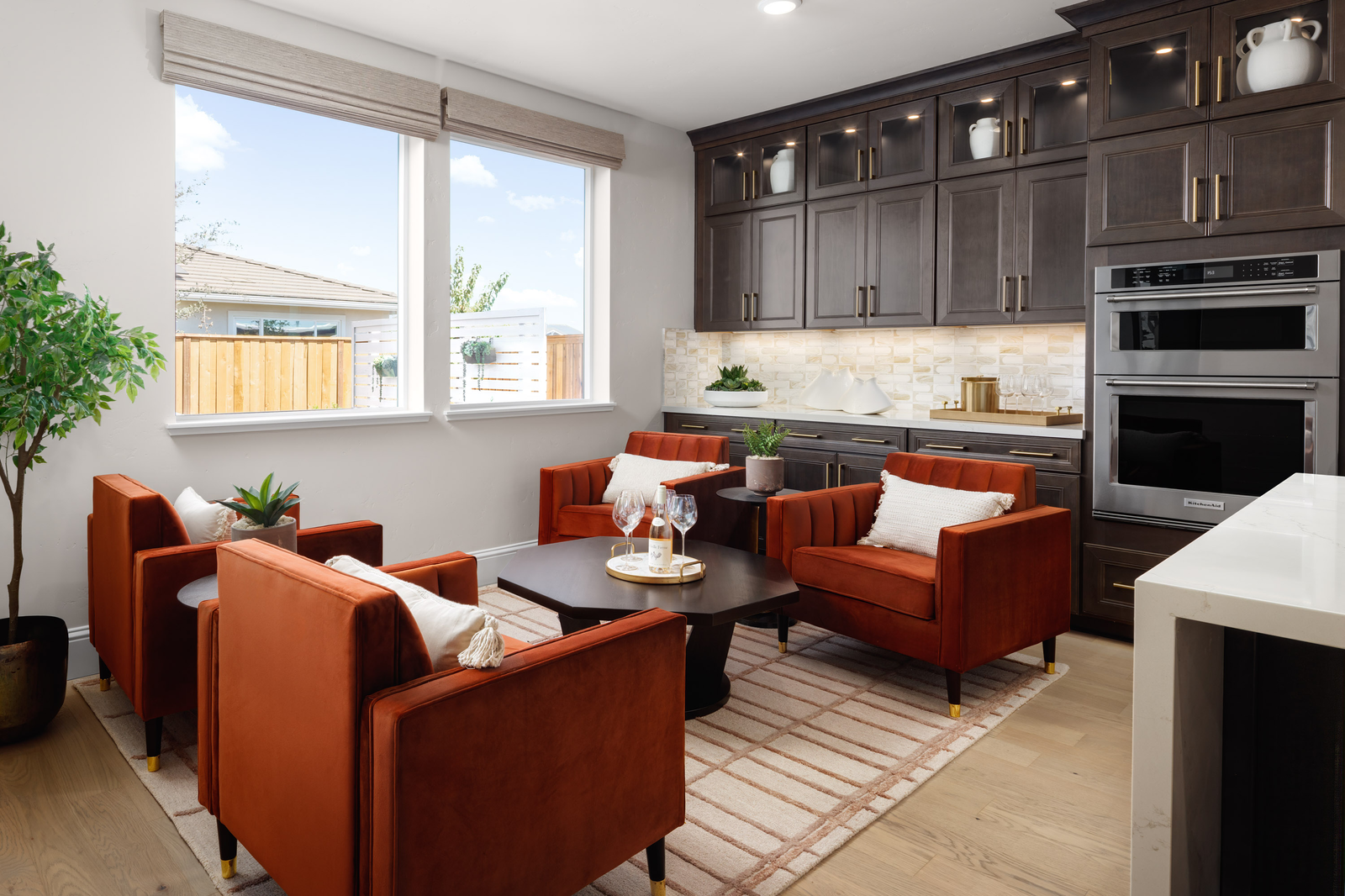 breakfast area with four rust-colored accent chairs, black coffee table with wine glasses, espresso cabinets, wood floors, and windows overlooking the backyard