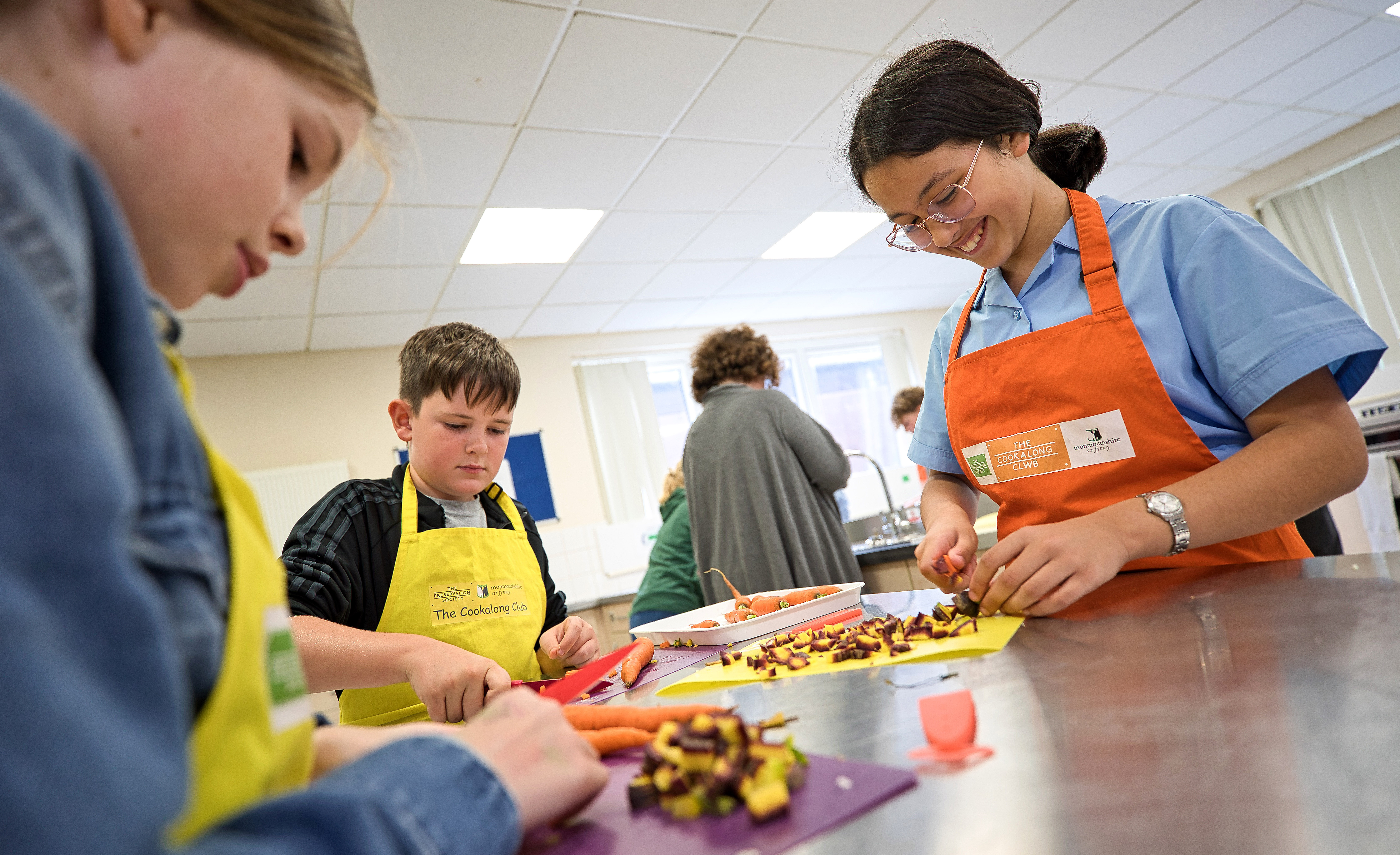 Three children wearing aprons happily chop vegetables on colorful cutting boards in a bright kitchen.