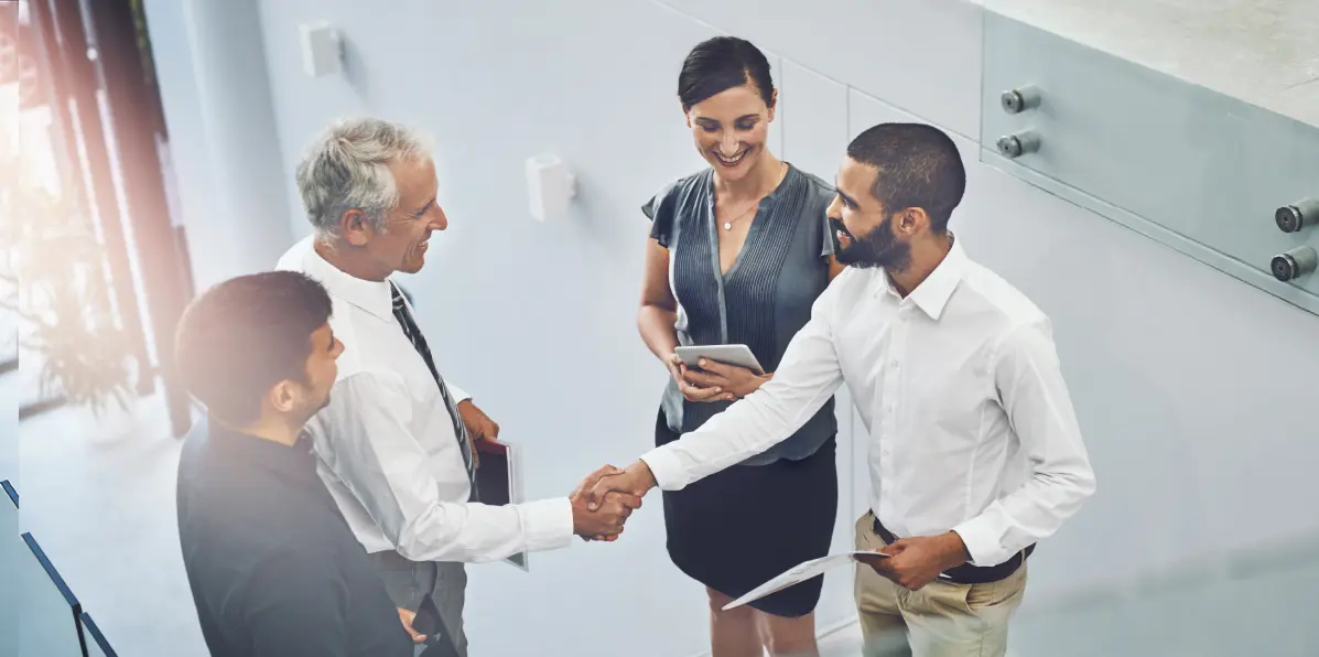 Four professionals engaged in a business meeting, with two individuals shaking hands while the others observe. The setting is modern, with bright lighting and a sleek, minimalistic office background.