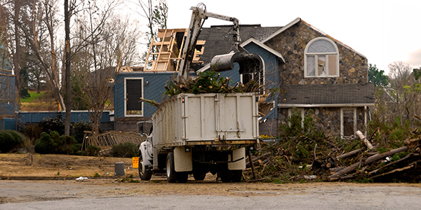 A partially damaged home made of wood and stone is surrounded by fallen trees. 