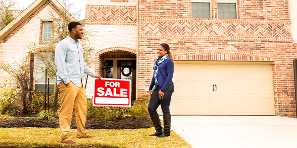 A couple standing by a 'For Sale' sign in front of a brick house.