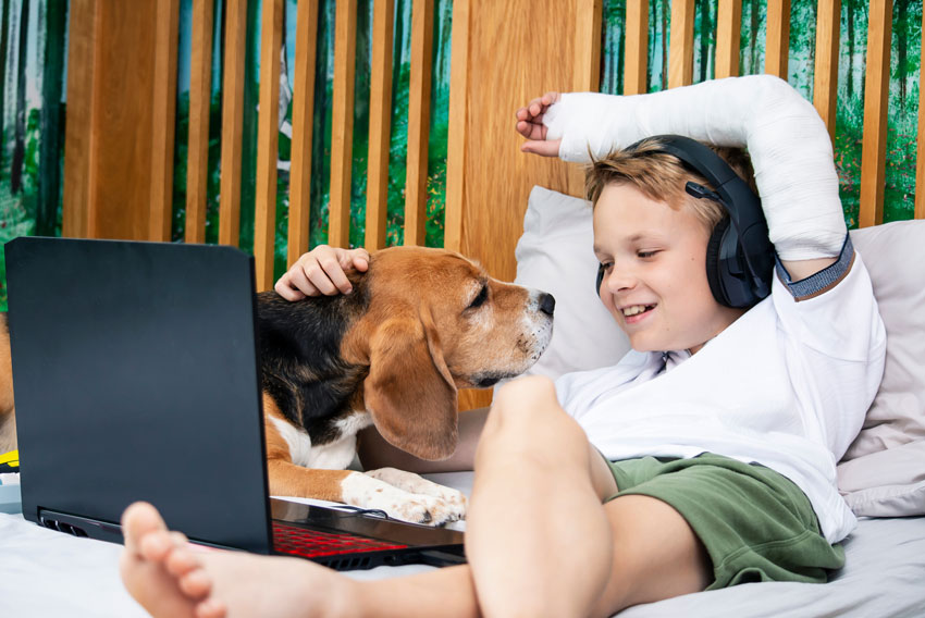 Deček, ki ima zaradi nezgode gips, s psom leži na postelji in gleda filme na računalniku.
