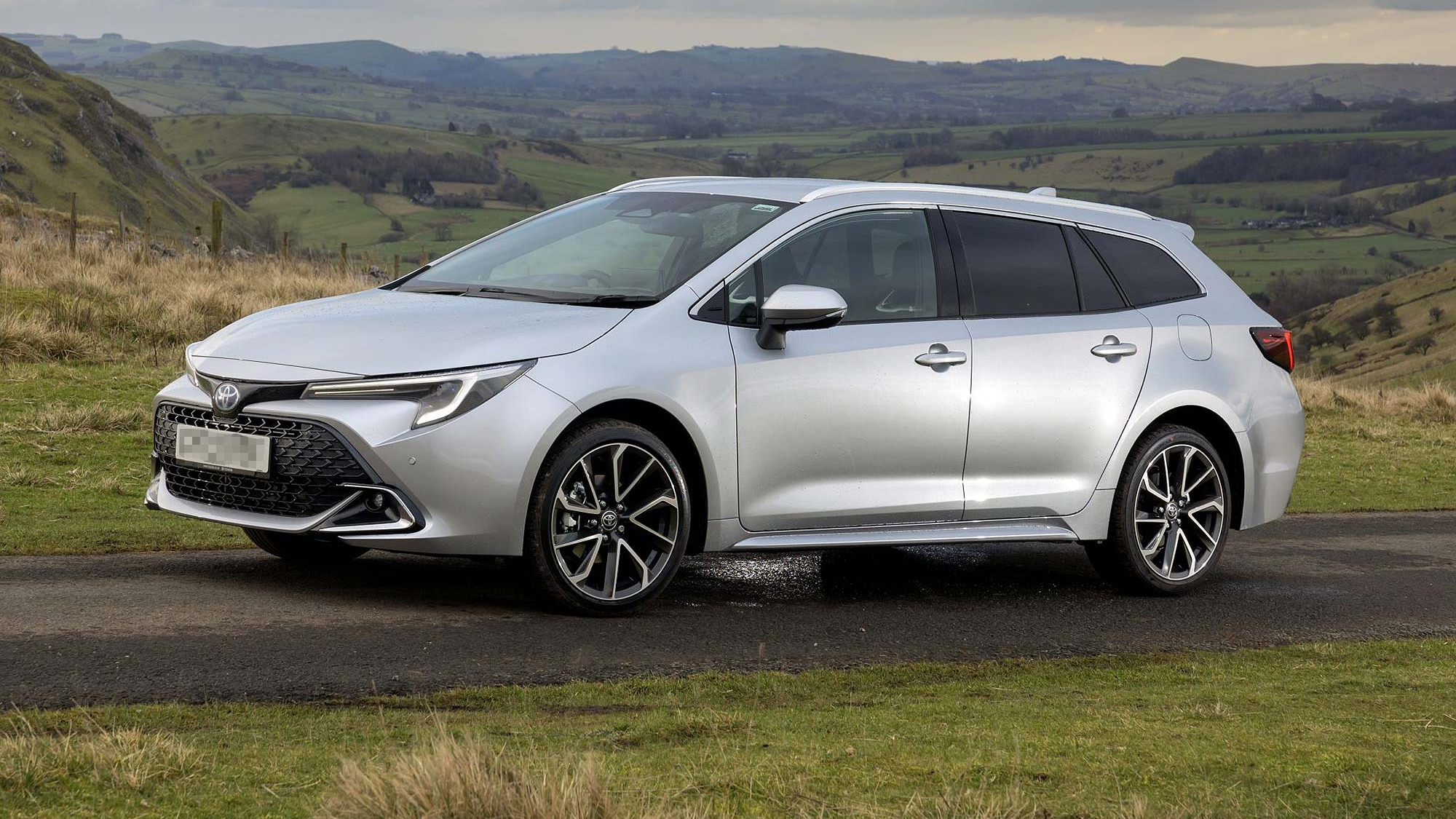Silver Toyota Corolla Touring Sports parked on a rural roadside, side and front three quarter view, with rolling hills and countryside in the background.