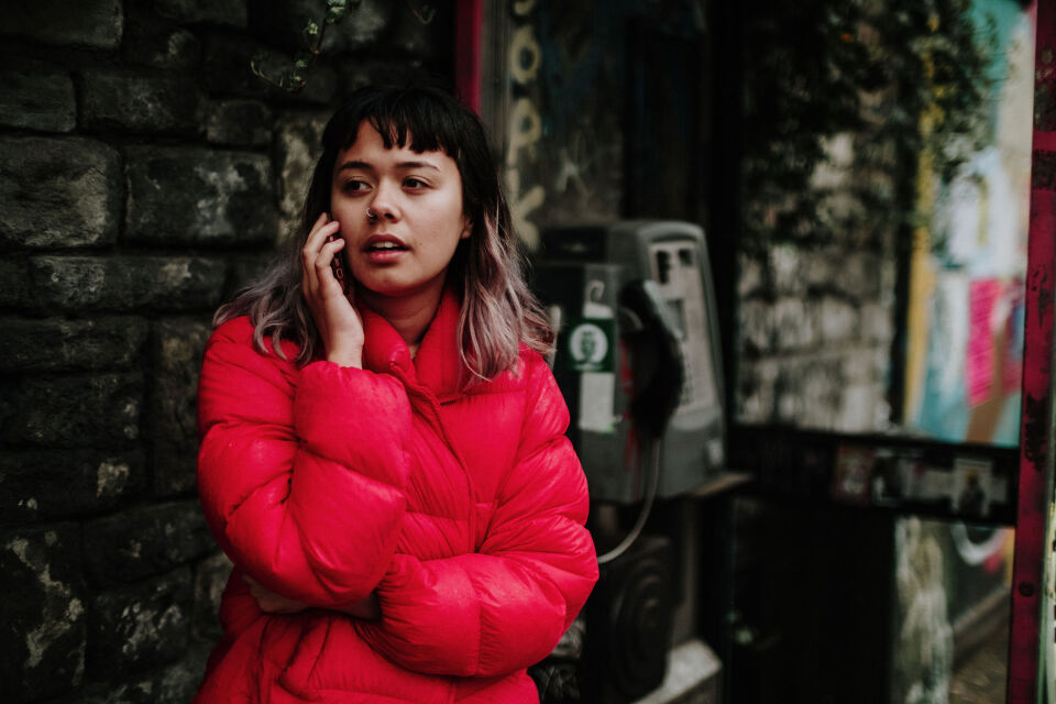 A young woman in a bright red puffer jacket talking on a mobile phone while leaning against a dark stone wall next to a public payphone.