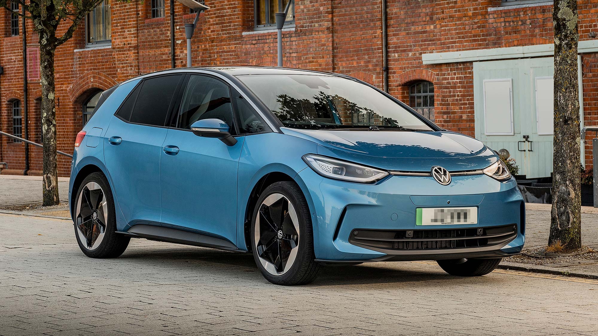 Blue Volkswagen ID.3 electric hatchback parked on a paved street in front of red brick buildings and trees.