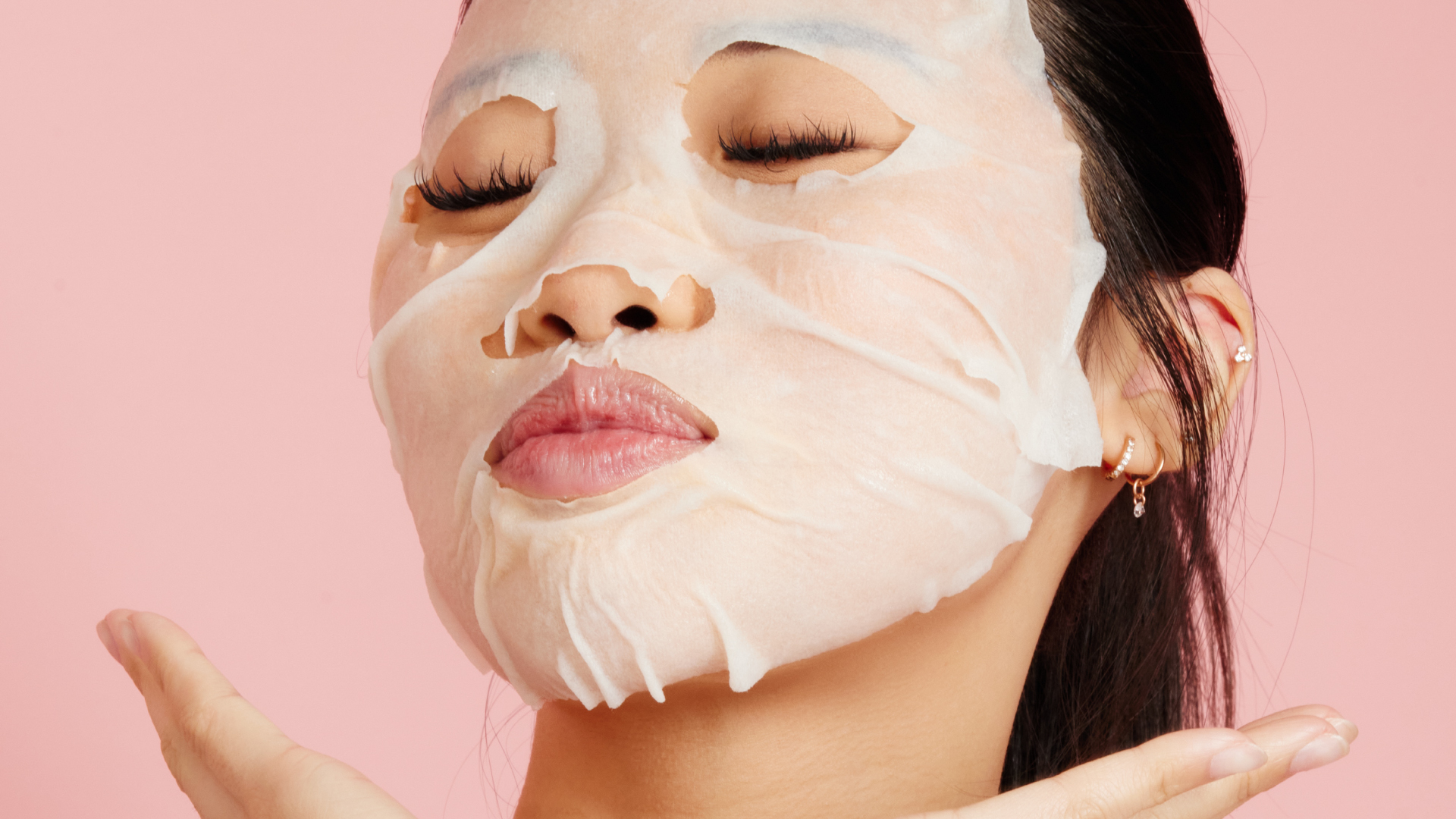 Woman wearing a hydrating H2O+ Vitamin C Glow Face Mask with eyes closed on a pink background.