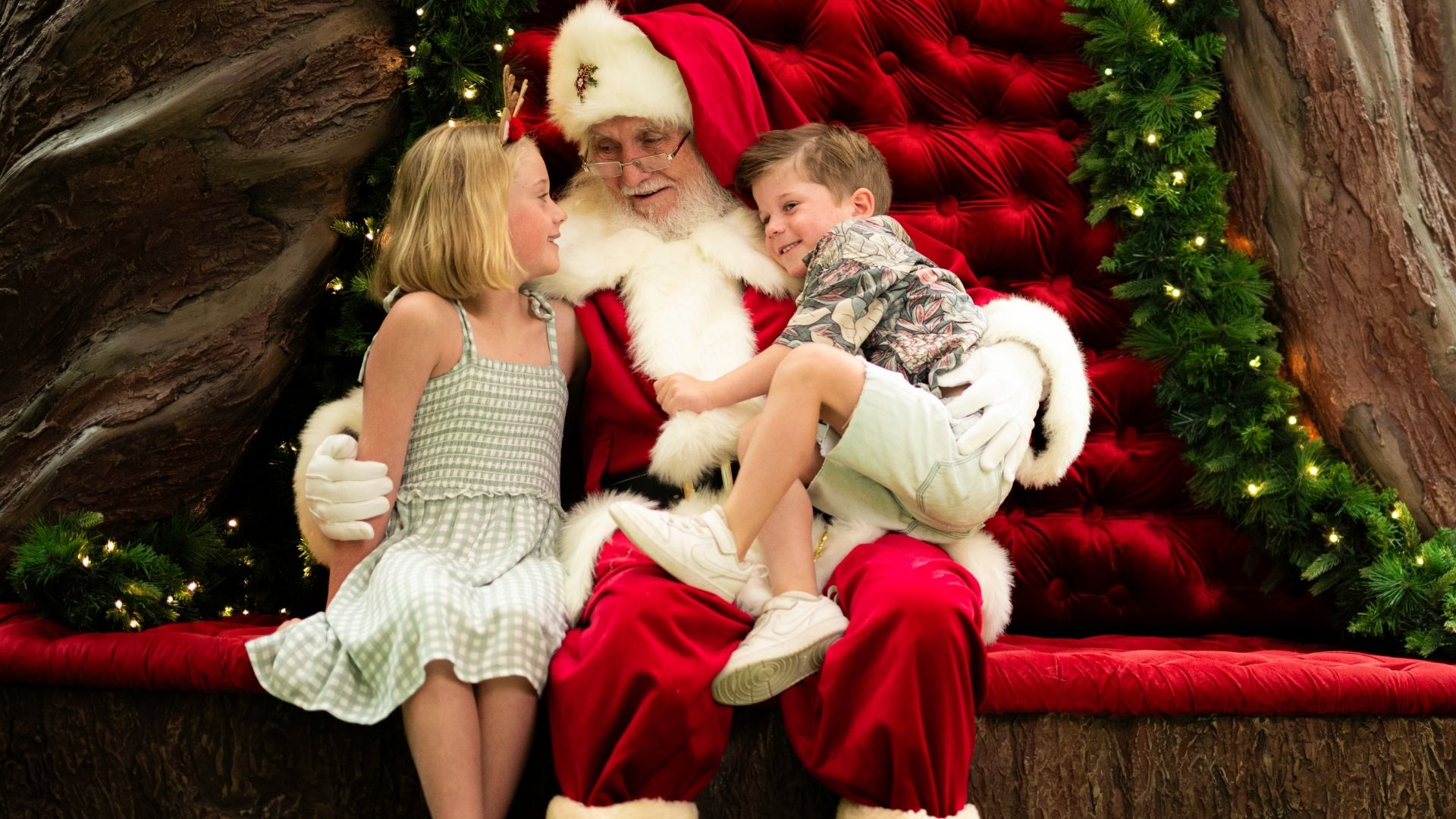 Two children sit with Santa on his red velvet throne, surrounded by lit Christmas garlands.