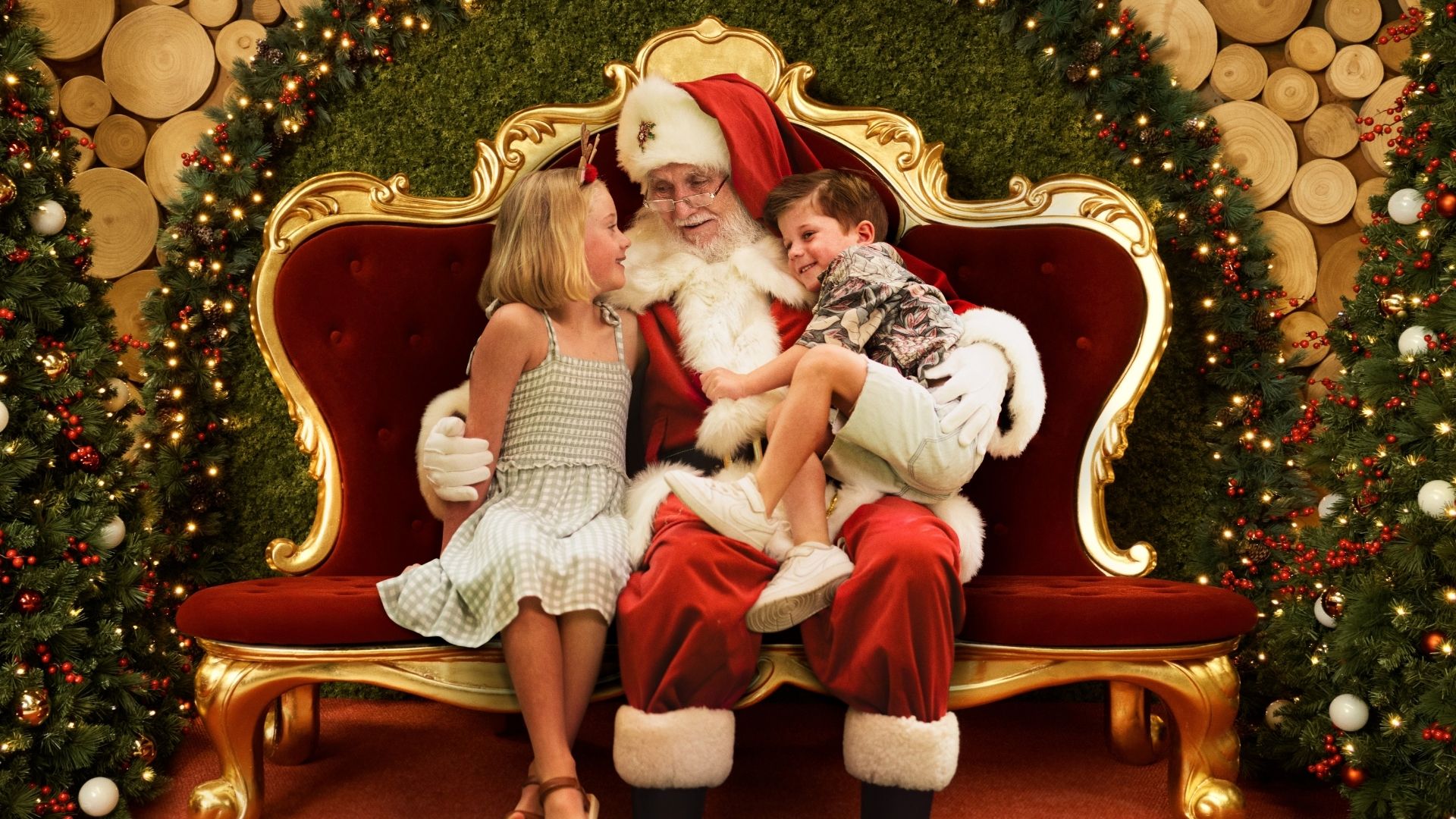 Two children sit with Santa on his red velvet throne, surrounded by lit Christmas garlands.