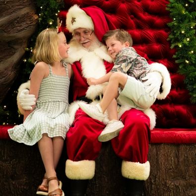 Two children sit with Santa on his red velvet throne, surrounded by lit Christmas garlands.