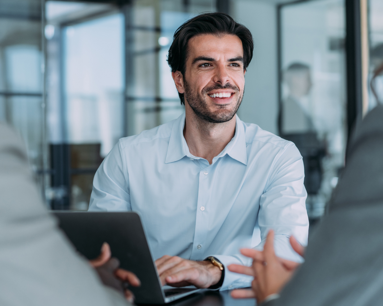 man in corporate attire with laptop in front of him on table speaking to others in office setting 