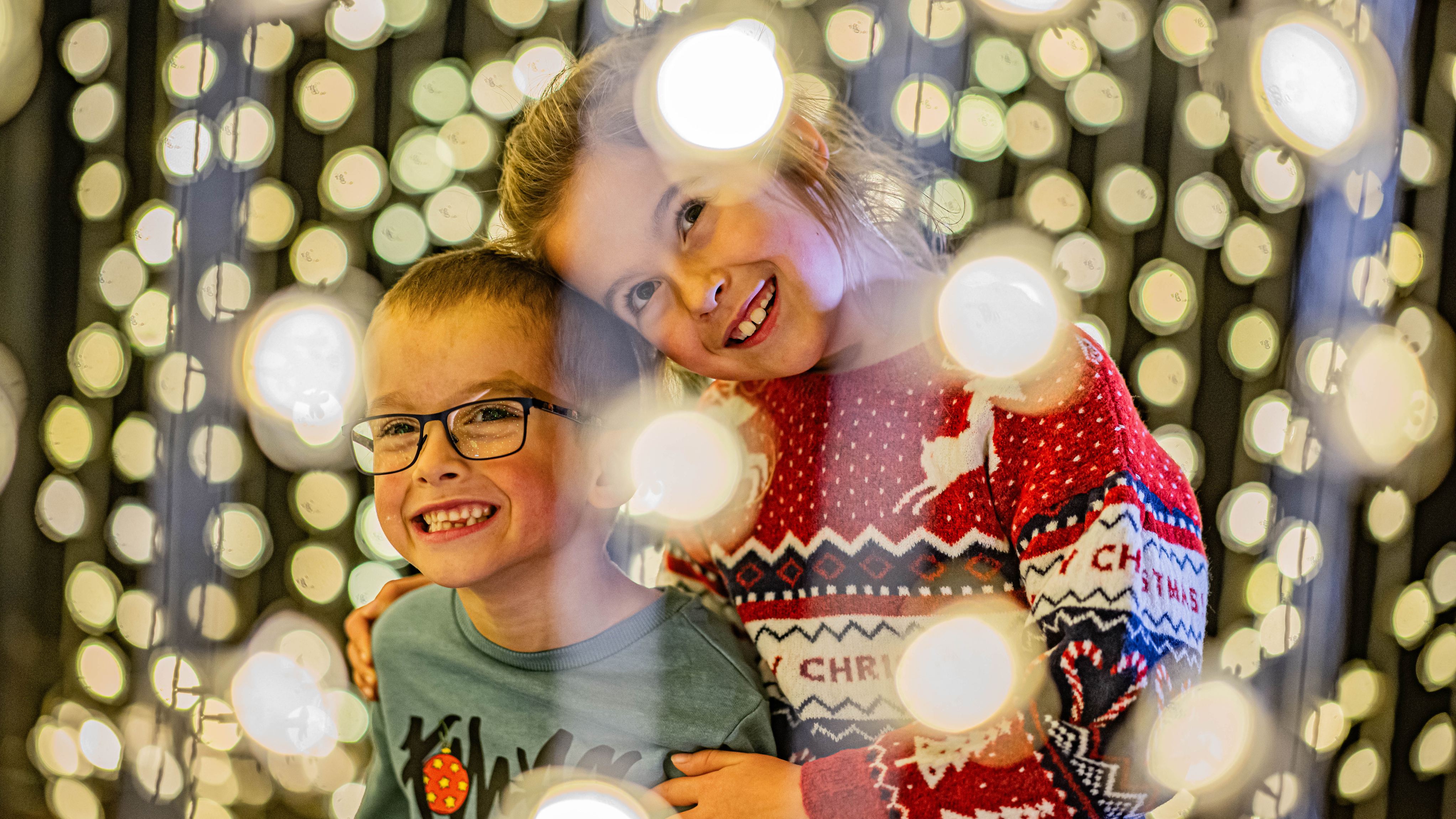 Two smiling children, one wearing glasses and the other a red Christmas jumper, pose together surrounded by glowing circular Christmas lights.