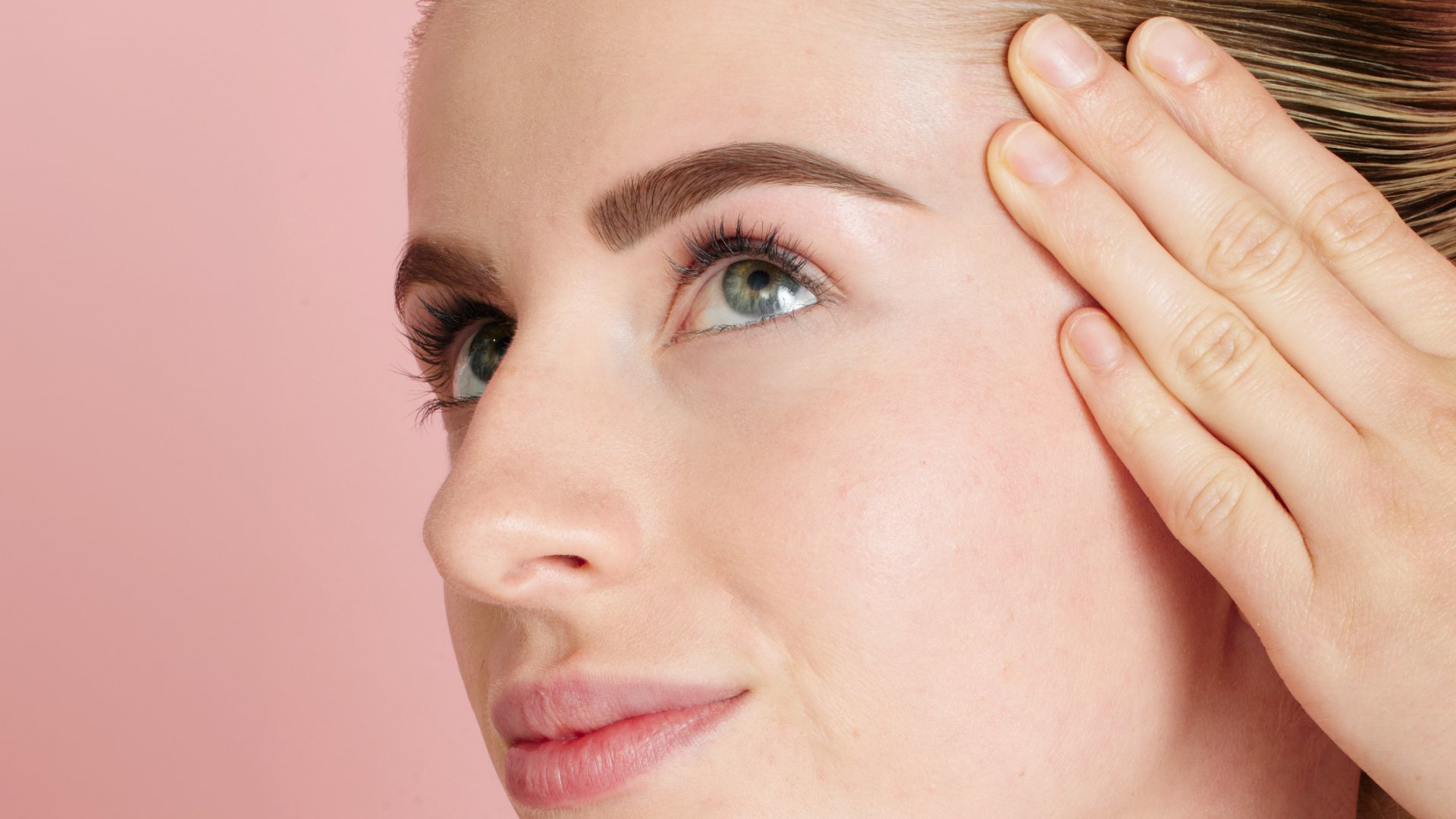 Close-up portrait of a woman with blonde hair and natural makeup, highlighting a neatly shaped and filled eyebrow, with her hand gently touching her temple against a soft pink background.