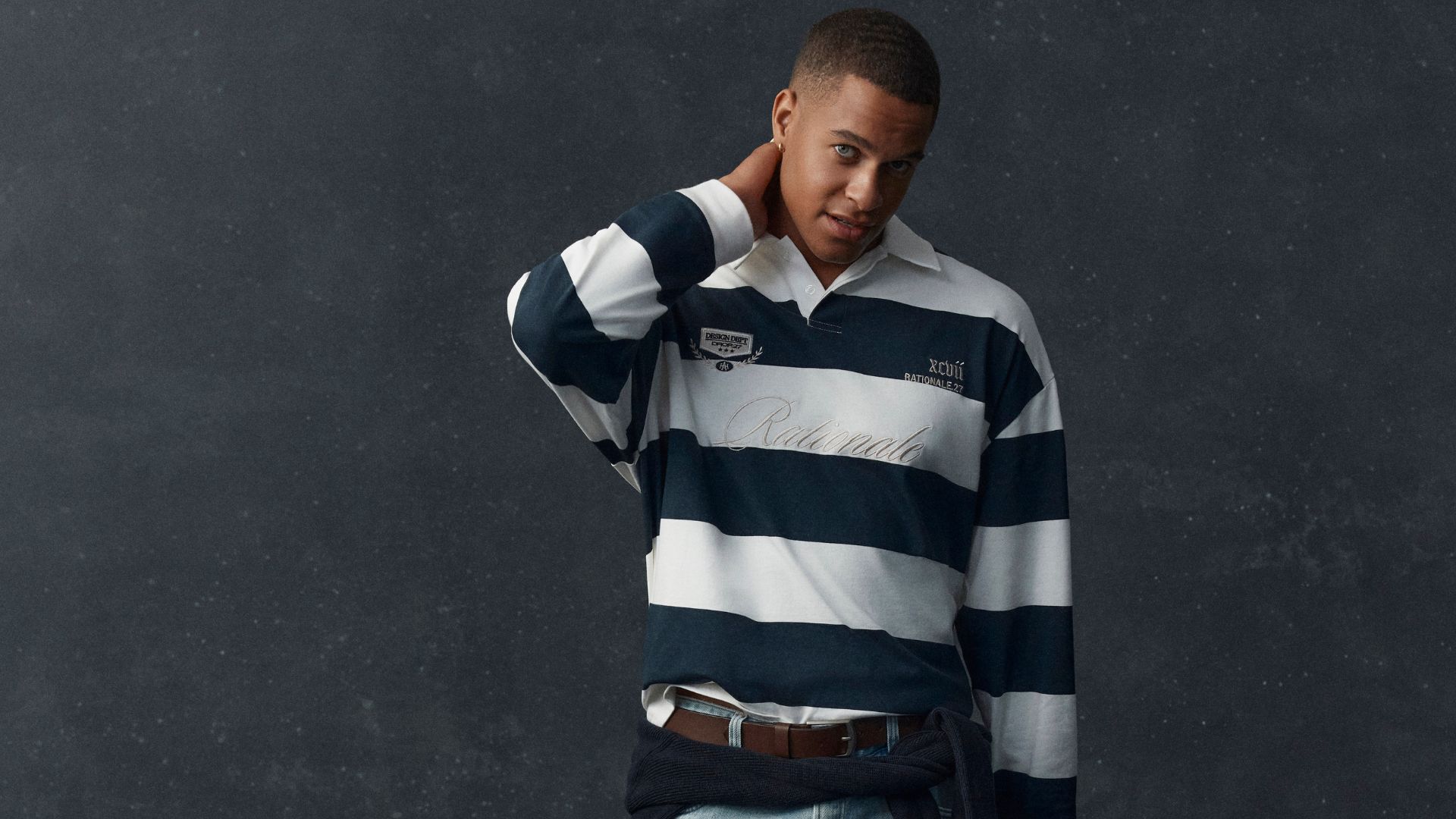 Model wearing a navy and white striped long sleeve rugby polo with light wash jeans, a brown belt and a navy knit tied around the waist.