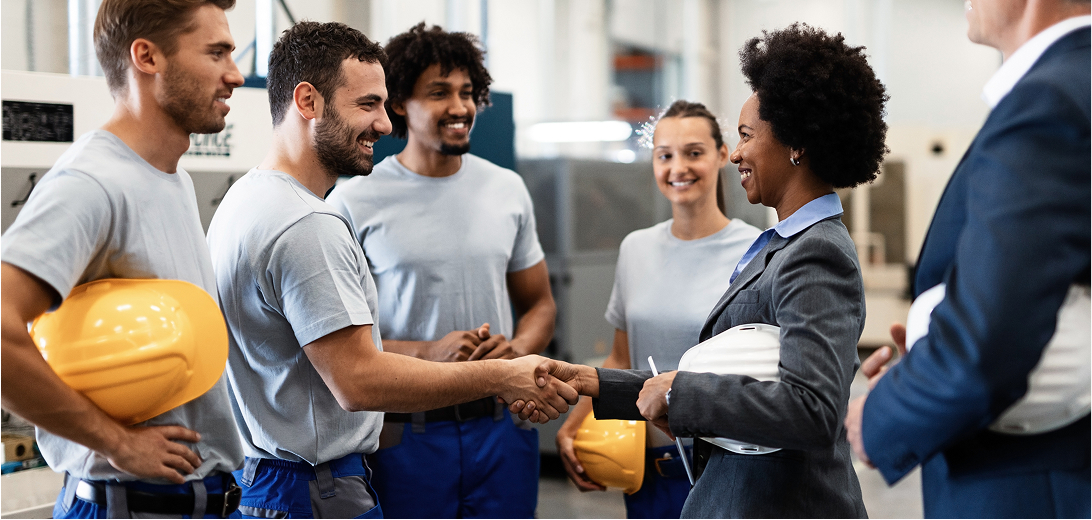 An indoor industrial or warehouse setting where a woman in a grey business suit is shaking hands with a male worker in a grey shirt and blue trousers. Three other workers and a man in a dark suit stand nearby, smiling. Several workers hold yellow or white safety helmets.