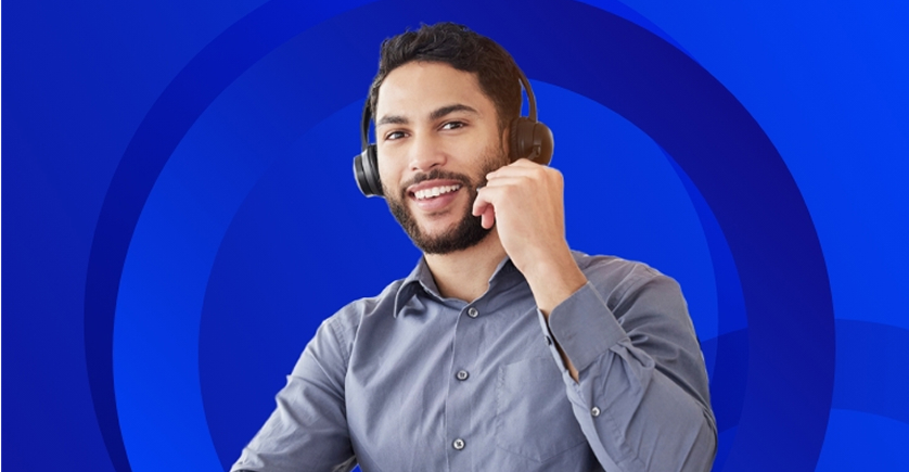 
The image shows a man wearing a headset with a microphone and a gray collared shirt.

He has dark hair and a beard, and he is smiling while looking forward. He appears to be working in a customer service or call center role, holding the headset's microphone near his mouth.