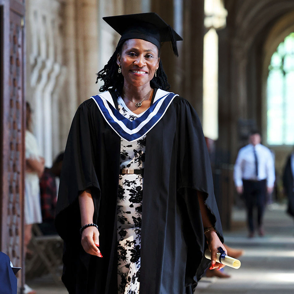 A smiling woman in a black graduation gown and cap, with a white and blue stole, walks down a cloistered hallway holding a rolled diploma.