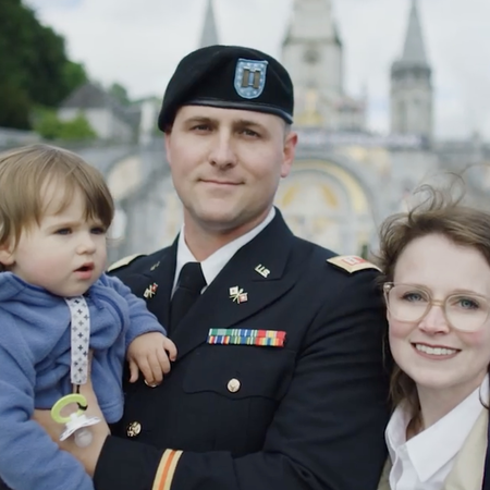 Woman stands with a military man in uniform holding a young baby girl with Lourdes France in the blurred background