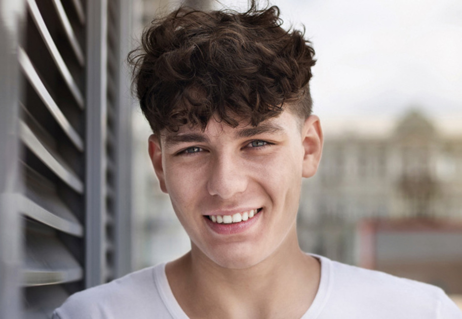 A young man with curly brown hair and light skin smiles at the camera outdoors, with blurred greenery and sunlight in the background. He is wearing a light-colored shirt.