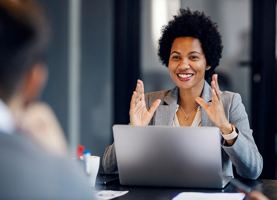 A smiling woman with short, curly dark hair sits at a conference table with a laptop open and speaks enthusiastically, gesturing with both hands. She is wearing a light gray suit jacket and a necklace. An out-of-focus person's back is visible across the table.