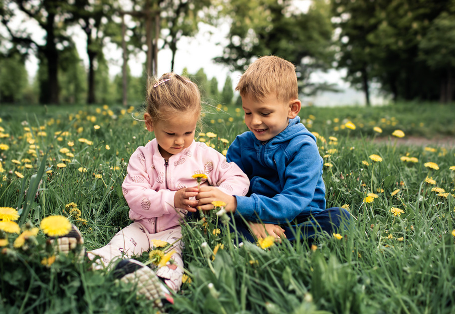 Two young children, a girl in a pink jacket and a boy in a blue hoodie, sit together in a grassy field with yellow dandelions, smiling and playing outdoors on a cloudy day. Trees are visible in the background.