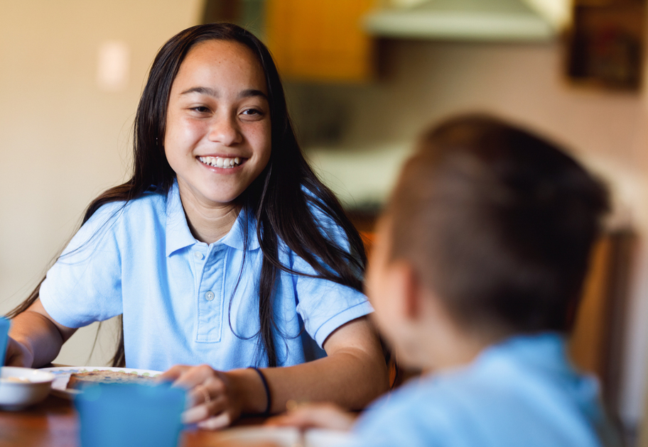 A girl with long dark hair, wearing a light blue polo shirt, smiles while sitting at a table with a younger child who is also in a blue shirt. They appear to be sharing a meal in a home setting.