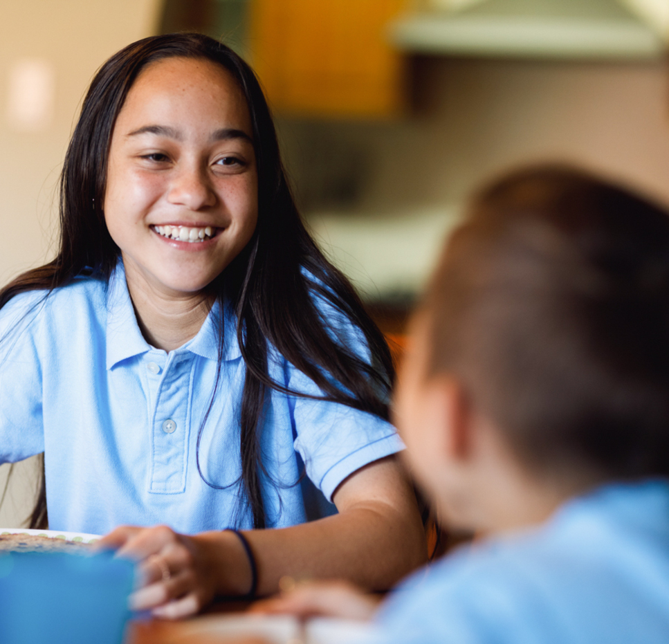 A girl with long dark hair, wearing a light blue polo shirt, smiles while sitting at a table with a younger child who is also in a blue shirt. They appear to be sharing a meal in a home setting.
