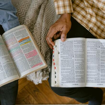 Two people sit on a couch, both are holding and looking at Bibles. 