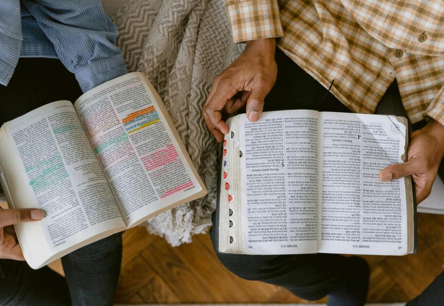 Two people sit on a couch, both are holding and looking at Bibles.