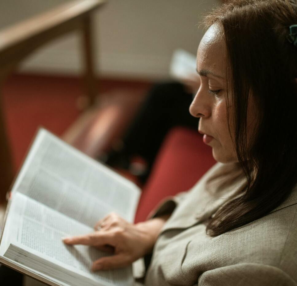 A woman with brown hair and a green hair clip sits on a wooden bench, reading the bible and pointing at a page with her finger. The setting appears to be quiet and indoors.