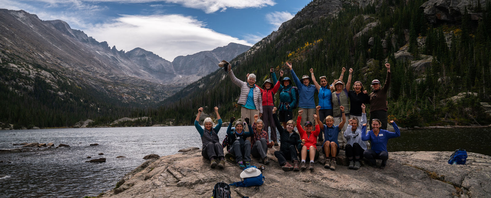 A group of women celebrate on a large rock by a mountain lake during a women-only walking and hiking trip.