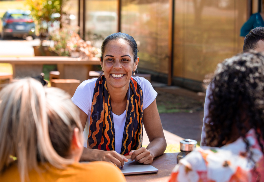	
A woman smiles while sitting at an outdoor table with three other people, enjoying a sunny day. She has a notebook in front of her and appears to be engaged in a friendly conversation.