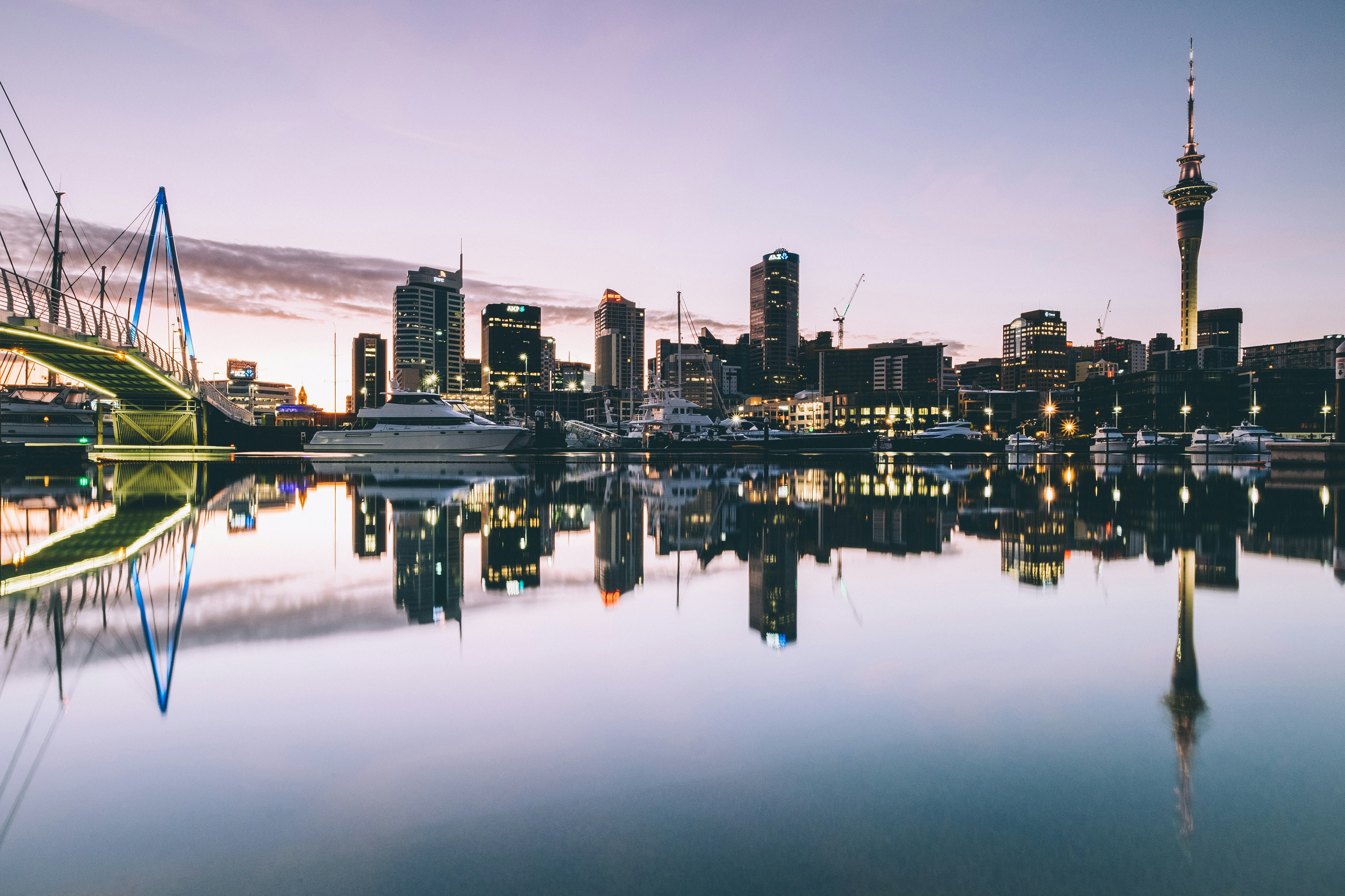 panoramic view of a modern cityscape at dusk or dawn, with the city's lights reflecting vividly on the calm, glassy surface of the water in the foreground. On the left, a cable-stayed bridge with illuminated blue and yellow accents connects to the city. The skyline features numerous high-rise buildings, some with glowing windows, and on the far right, a distinctive tall observation tower (likely the Sky Tower in Auckland) stands prominently against the soft, pastel-colored sky. Several boats and yachts are docked along the waterfront, adding to the urban marina scene.