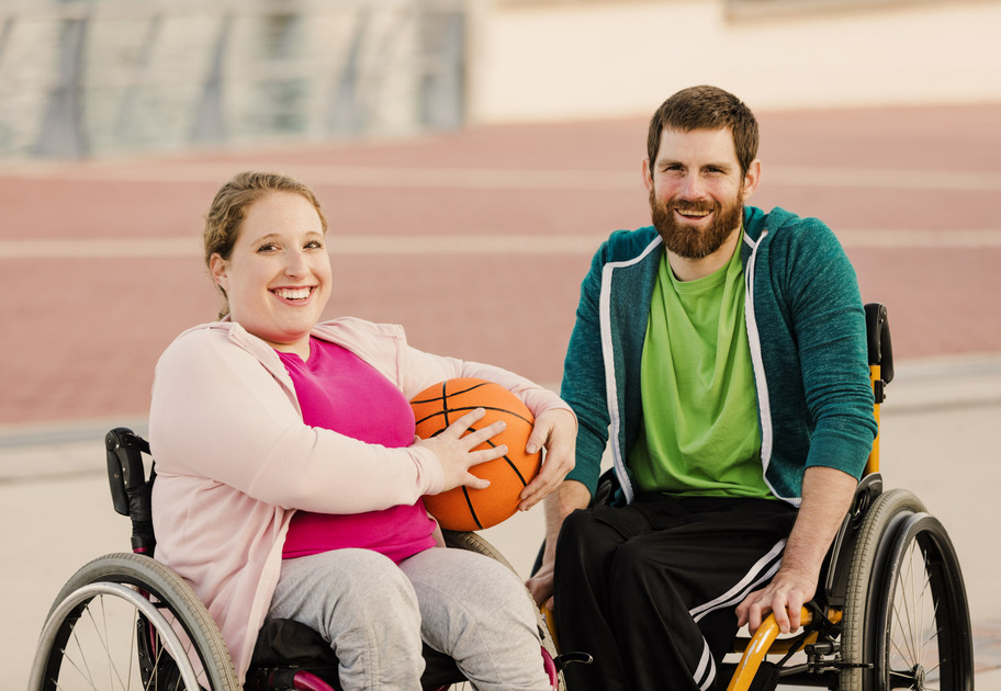 A young woman and a young man outdoors playing basketball. The woman is a wheelchair user, and both are smiling toward the camera