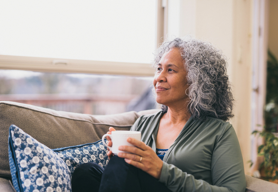 An older woman with curly gray hair sits on a couch by a window, holding a white mug and looking thoughtfully outside. She is relaxed, surrounded by blue patterned pillows, with natural light filling the room.
