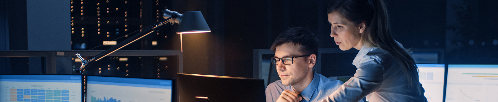 A man and a woman are working together at computer monitors in a dark office or server room, lit by a desk lamp. The woman is leaning over the man's shoulder to look at his screen.