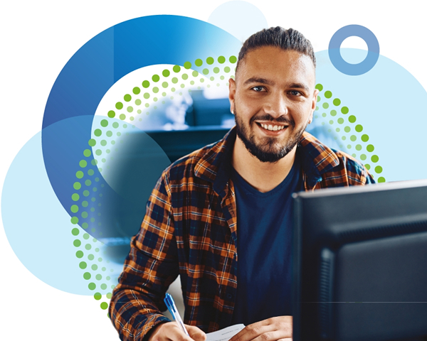 A smiling man with a beard and dark hair tied back sits at a desk, looking at the camera while taking notes. He is wearing a blue t-shirt and a plaid shirt, surrounded by a light-blue and dotted green and yellow circular graphic overlay.