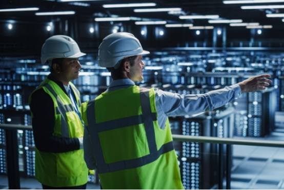 Two men in hard hats and high-visibility vests stand in a large server room, with one pointing into the distance.