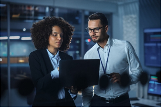 A man and a woman in business attire look at a laptop screen together in a modern, dark office environment.