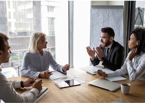 Four professionals—two men and two women—are seated around a large wooden conference table in a sunlit room, engaged in a discussion. A man in a suit gestures with his hands while speaking to a woman across from him.