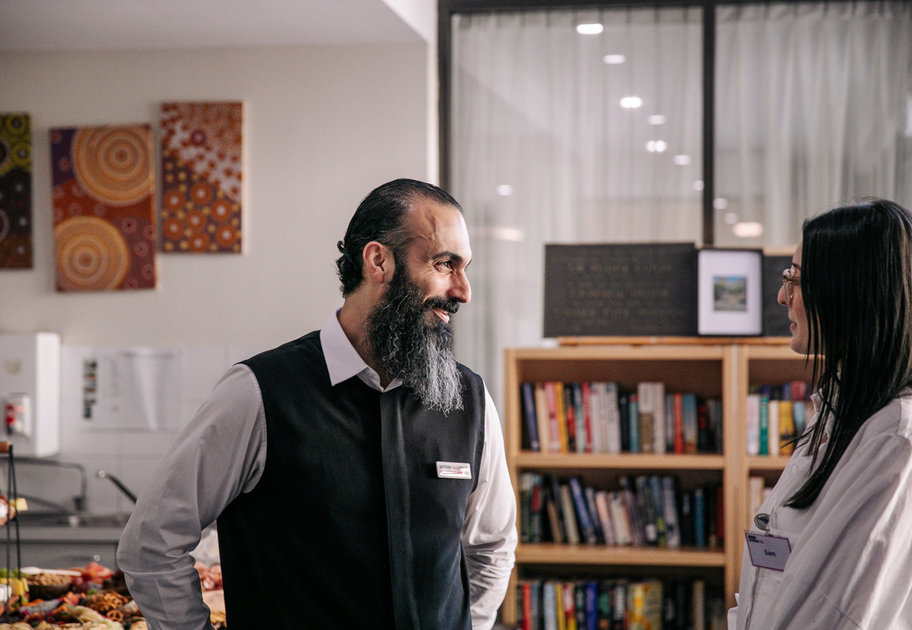 A man with a beard wearing a black vest and name tag smiles while talking to a woman in glasses and a white coat, standing in a room with bookshelves, food, and colorful artwork on the wall.