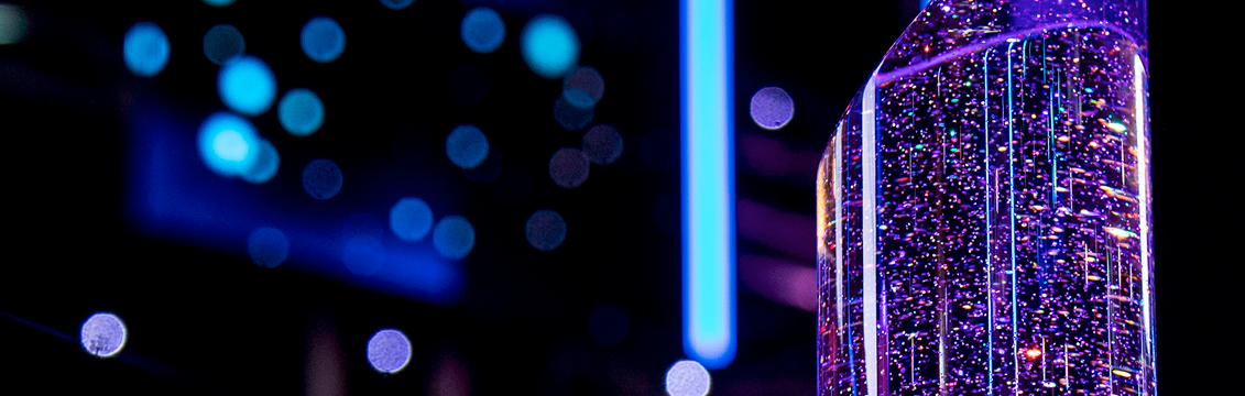 An abstract, dark image featuring a close-up of a decorative, cylindrical object filled with bubbles, lit up in vibrant purple and blue light. Out-of-focus blue lights and a vertical bright blue light strip are visible in the background, suggesting an awards ceremony atmosphere.