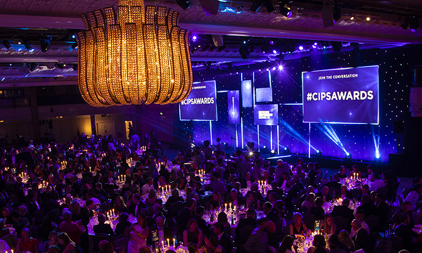 A wide-angle view of a large, formal awards dinner or gala event. Attendees are seated at round tables lit by candlelight. In the foreground, a massive, ornate chandelier hangs from the ceiling. On the stage in the background, bright spotlights illuminate large screens displaying the hashtag #CIPSawards against a backdrop of blue and purple lighting.