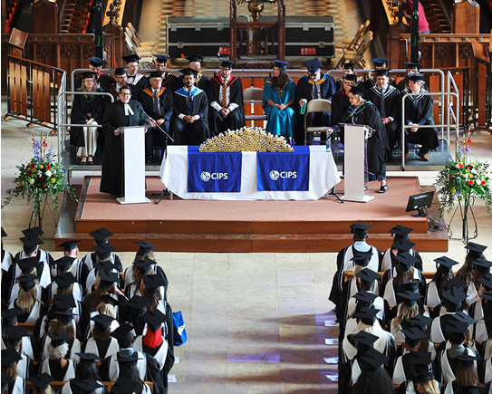 An image from a graduation ceremony taking place inside a large hall, showing many graduates in caps and gowns seated in the foreground, and officials or speakers on a raised platform in the background behind a table with two CIPS logos.