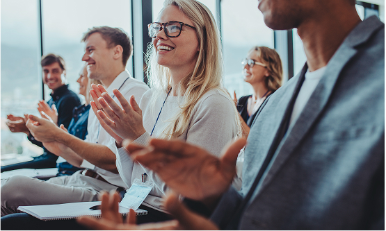 A group of people sitting in a sunlit venue, clapping enthusiastically. A blonde woman with glasses in the foreground is smiling widely and clapping.